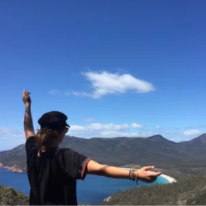 Eine Frau blickt von oben mit erhobenem Arm auf eine weite Landschaft mit blauem Meer und Bergen während einer Auslandsreise.