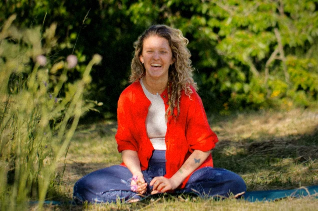 Yogalehrerin und Ergotherapeutin Madeleine Zimmer sitzt lachend im Schneidersitz auf einer Yogamatte in der Natur.