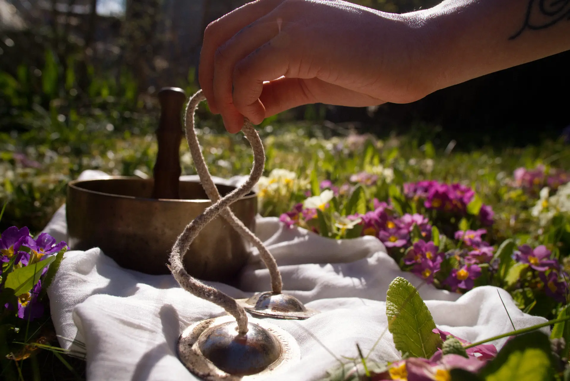 Eine Hand hält Zimbeln (Tingshas) über einer tibetischen Klangschale auf einer Frühlingswiese als Banner für Sonnenkind Yoga.